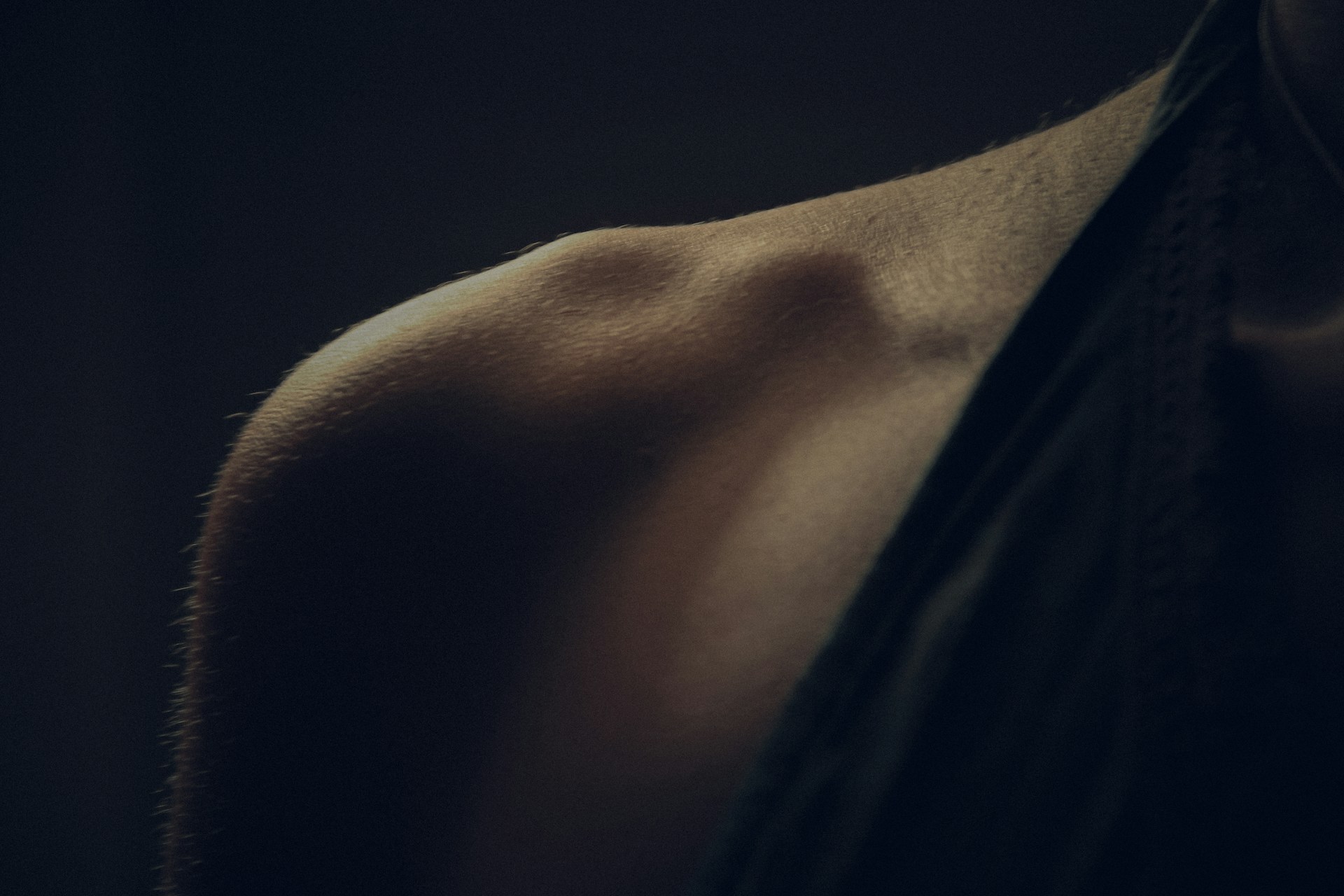A close-up of a woman's exposed neckline and collarbone softly illuminated by moody, golden light against a velvet dark background evoking intimacy and mystery.