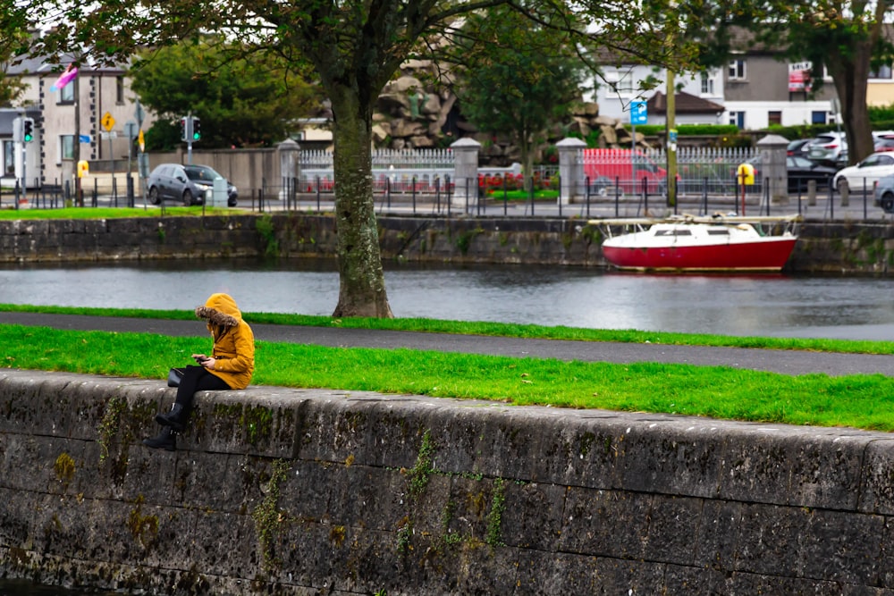 man in yellow jacket sitting on concrete bench near red and white boat during daytime