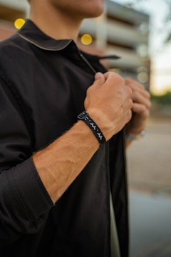 A person is adjusting a black jacket while wearing a black bracelet with white designs on their wrist. The background is slightly blurred, showing what appears to be an urban setting with lights.