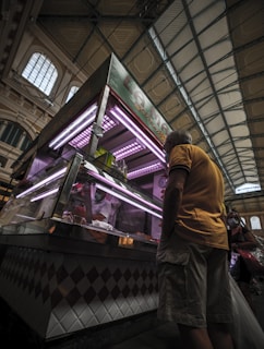 A dimly lit indoor market scene with a man in a yellow shirt standing in front of a meat counter. The counter features glass panes and is illuminated by purple lights. Behind the counter, a worker wearing a mask is visible, handling products. The market has an industrial vibe with its high ceilings and large windows.