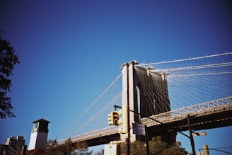 A large suspension bridge with thick cables spans across the sky, featuring a tall stone tower with arches. In the foreground, there are street signs and a traffic light. The blue sky contrasts with the structure's grey tones, while trees are visible near the base of the bridge.