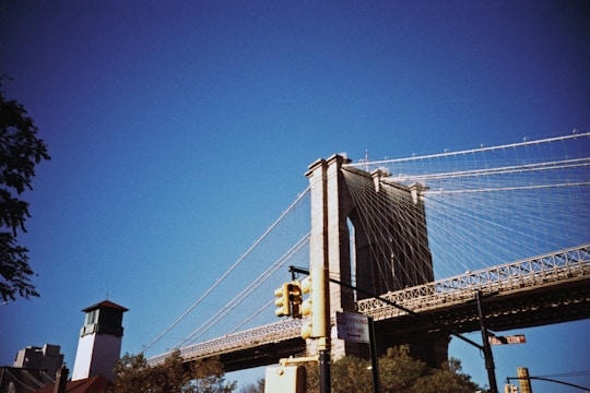 A large suspension bridge with thick cables spans across the sky, featuring a tall stone tower with arches. In the foreground, there are street signs and a traffic light. The blue sky contrasts with the structure's grey tones, while trees are visible near the base of the bridge.