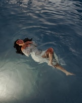 A woman in a white dress floats peacefully in a body of water. Her eyes are closed, and her hands are gently placed on her chest. The water around her is calm, with gentle ripples reflecting the light.