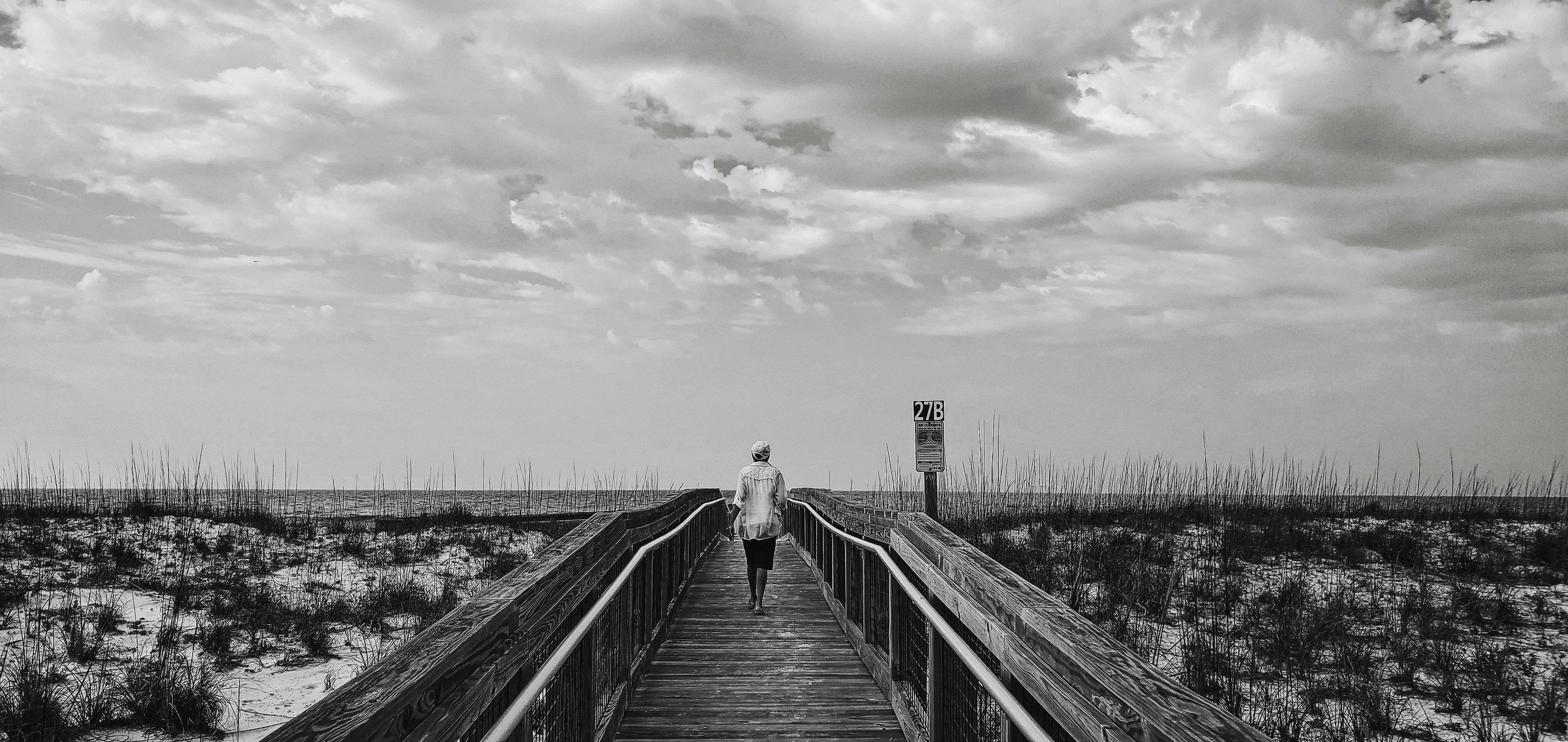 grayscale photo of man and woman walking on wooden bridge