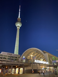 A nighttime city scene featuring a tall television tower with a brightly lit spire. The foreground shows a modern glass and steel structure with the illuminated sign 'Alexanderplatz'. The sky is clear and dark, enhancing the prominence of the tower.