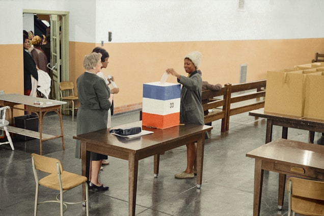 A woman is casting her vote into a ballot box that is painted with stripes of blue, white, and red. She is surrounded by two other people who seem to be assisting or monitoring the voting process. The room is large with a simple, clean design, featuring several tables and chairs. There are additional boxes and some paper materials on the tables.