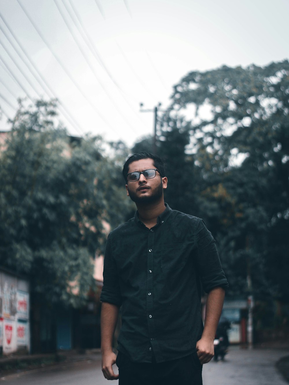 man in black button up shirt wearing black sunglasses standing near green trees during daytime