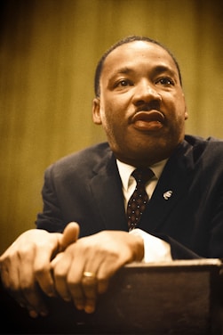 A man in a suit is leaning forward with clasped hands resting on a surface. He appears thoughtful and attentive, wearing a dark suit with a patterned tie and a small pin on his lapel. The background is softly blurred with warm tones.