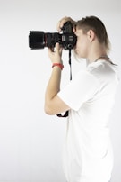 A person is holding a camera up to their eye, taking a photo. They are wearing a white t-shirt and have light-colored hair. The setting is minimalistic with a plain white background, emphasizing the action of photography.