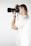 A person is holding a camera up to their eye, taking a photo. They are wearing a white t-shirt and have light-colored hair. The setting is minimalistic with a plain white background, emphasizing the action of photography.