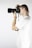 A person is holding a camera up to their eye, taking a photo. They are wearing a white t-shirt and have light-colored hair. The setting is minimalistic with a plain white background, emphasizing the action of photography.