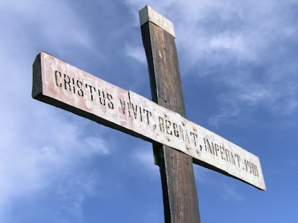 A tall wooden cross stands against a backdrop of a bright blue sky with scattered clouds. The cross has an inscription in Latin that reads 'CRISTUS VIVIT, REGNAT, IMPERAT. 1934'. The sun's illumination highlights the grain of the wood and the texture of the painted inscription.