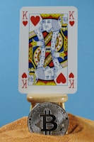 A playing card with a king of hearts design is placed on a small wooden stand. Below the card, a silver Bitcoin coin rests on a mound of sand, set against a light blue background.