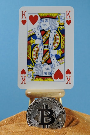 A playing card with a king of hearts design is placed on a small wooden stand. Below the card, a silver Bitcoin coin rests on a mound of sand, set against a light blue background.