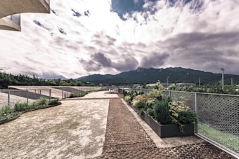 A modern architectural space featuring a rooftop garden with green plants and a pathway bordered by metal fencing. The sky is cloud-covered, with dramatic clouds casting shadows over distant mountains.