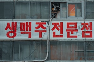 A building facade features large red Korean letters on a white banner sign. Above, weathered windows show signs of neglect with cracked glass and exposed wiring. The overall appearance suggests age and urban decay.