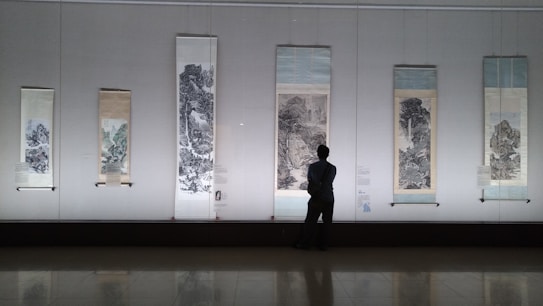 Several traditional scroll paintings are displayed in a gallery, each depicting intricate landscapes. A person is standing in front of the paintings, observing them thoughtfully.