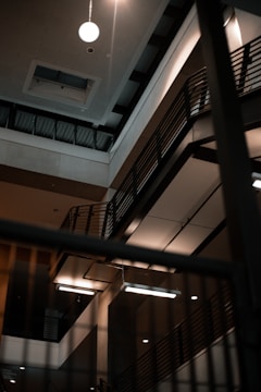 Interior of a multi-story building featuring modern architecture with metal railings and exposed beams. The lighting is subdued, with a few bright fixtures illuminating the area.