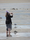 A person standing barefoot on a wet sandy beach, wearing a black top, grey skirt, and purple headscarf, taking a picture with a smartphone. There are a few gulls nearby and a pair of black slip-on shoes placed on the sand.