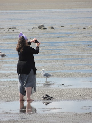 A person standing barefoot on a wet sandy beach, wearing a black top, grey skirt, and purple headscarf, taking a picture with a smartphone. There are a few gulls nearby and a pair of black slip-on shoes placed on the sand.