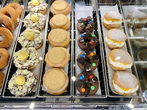 An assortment of doughnuts is displayed in a bakery case. On the left, there are plain sugar-coated doughnuts and those topped with white chocolate shavings and yellow buttons. The middle section showcases chocolate-covered doughnuts garnished with colorful candies and pieces of Oreo cookies. On the right, cream-filled doughnuts are dusted with powdered sugar.