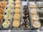 An assortment of doughnuts is displayed in a bakery case. On the left, there are plain sugar-coated doughnuts and those topped with white chocolate shavings and yellow buttons. The middle section showcases chocolate-covered doughnuts garnished with colorful candies and pieces of Oreo cookies. On the right, cream-filled doughnuts are dusted with powdered sugar.