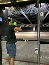 A person wearing ear protection is aiming a firearm at a shooting range. The shooting lane is long, extending to a target hung at the far end. The floor is scattered with bullet casings, and the lighting above is bright, illuminating the range.