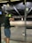 A person wearing ear protection is aiming a firearm at a shooting range. The shooting lane is long, extending to a target hung at the far end. The floor is scattered with bullet casings, and the lighting above is bright, illuminating the range.