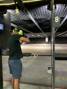 A person wearing ear protection is aiming a firearm at a shooting range. The shooting lane is long, extending to a target hung at the far end. The floor is scattered with bullet casings, and the lighting above is bright, illuminating the range.