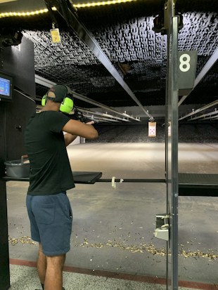 A person wearing ear protection is aiming a firearm at a shooting range. The shooting lane is long, extending to a target hung at the far end. The floor is scattered with bullet casings, and the lighting above is bright, illuminating the range.