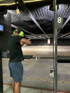 A person wearing ear protection is aiming a firearm at a shooting range. The shooting lane is long, extending to a target hung at the far end. The floor is scattered with bullet casings, and the lighting above is bright, illuminating the range.