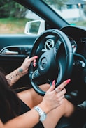 A person with long hair and tattoos on their arm is holding the steering wheel of a luxury car. The car features a prominent logo in the center of the steering wheel. The person's fingernails are painted bright red, and they are wearing a watch and rings. The interior of the car is sleek, with a side mirror visible through the window.