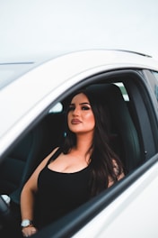 A woman with long, dark hair and makeup is sitting confidently in the driver’s seat of a white car, wearing a black sleeveless top and a watch on her left wrist. The focus is on her expression and style.