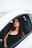 A woman with long, dark hair and makeup is sitting confidently in the driver’s seat of a white car, wearing a black sleeveless top and a watch on her left wrist. The focus is on her expression and style.
