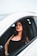 A woman with long, dark hair and makeup is sitting confidently in the driver’s seat of a white car, wearing a black sleeveless top and a watch on her left wrist. The focus is on her expression and style.