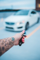 A hand with a tattoo holds a car key fob featuring a Mercedes logo, with a blurred white car in the background. The hand has bright pink nail polish, and the setting appears to be an outdoor parking area.