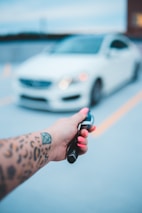A hand with a tattoo holds a car key fob featuring a Mercedes logo, with a blurred white car in the background. The hand has bright pink nail polish, and the setting appears to be an outdoor parking area.