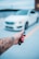 A hand with a tattoo holds a car key fob featuring a Mercedes logo, with a blurred white car in the background. The hand has bright pink nail polish, and the setting appears to be an outdoor parking area.