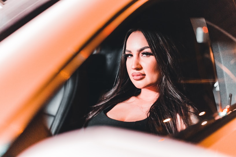 A person with long dark hair and vivid makeup is sitting inside a vehicle. The lighting highlights their features and creates a warm glow, with reflections visible on the window.
