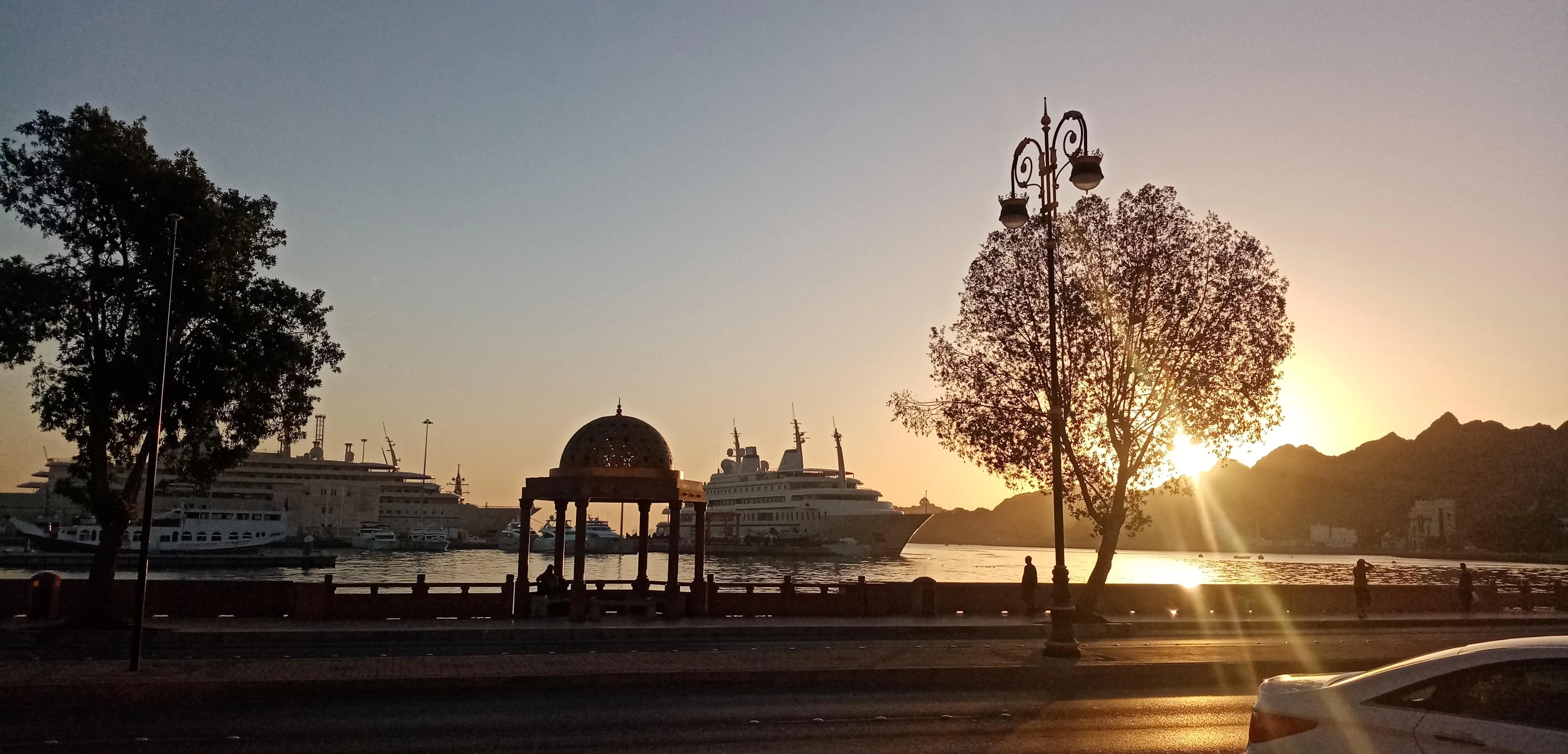 Silhouetted domed pavilion and moored ships along Muscat's corniche at sunset, with a flowering tree in the foreground.