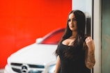 A woman with long dark hair is standing next to a white car, likely a luxury model, with a bright red background. She has a tattooed arm and is wearing a black outfit, exuding a confident and bold presence.
