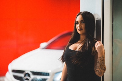 A woman with long dark hair is standing next to a white car, likely a luxury model, with a bright red background. She has a tattooed arm and is wearing a black outfit, exuding a confident and bold presence.