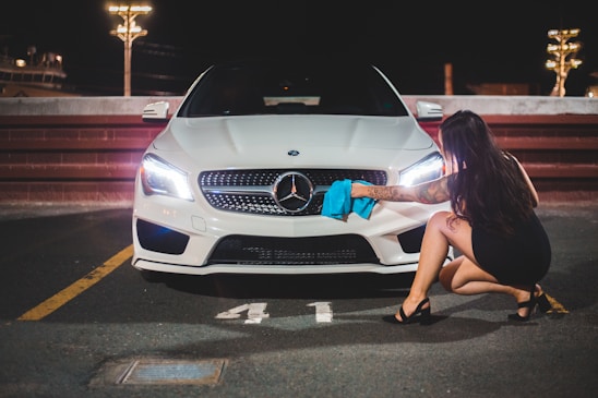 A woman in a black dress and heels is crouching next to a white Mercedes-Benz, using a blue cloth to clean the front of the car in a dimly lit parking area at night.