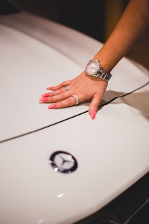 A hand with pink manicured nails rests on a car hood adorned with a luxury car emblem. The wrist is adorned with a metallic watch and rings on multiple fingers.
