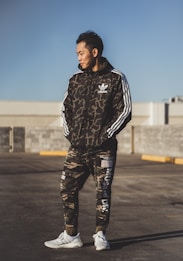 A person stands outdoors wearing a camouflage-patterned athletic outfit with the logo of a well-known sports brand. The individual appears to be in a parking lot or open area under clear daylight. The outfit consists of a jacket with white stripes on the sleeves and matching pants, paired with white sneakers.