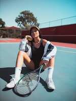 A person sitting on a tennis court, wearing a visor, black sports outfit, and white sneakers. They hold a tennis racket and have a tennis ball under their chin. The court is outdoor and sunny with a fence and trees in the background.