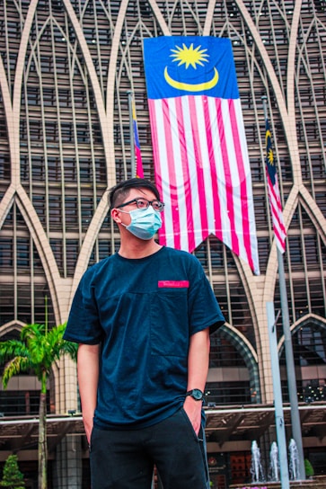 A person wearing a face mask and glasses stands in front of a building with an intricate architectural design. The Malaysian flag is prominently displayed behind the person, along with other small flags.