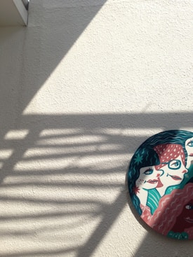 An artistic round piece placed on a textured wall, featuring stylized faces in vibrant colors. Shadows from a grid pattern are cast across the wall, adding depth to the composition.