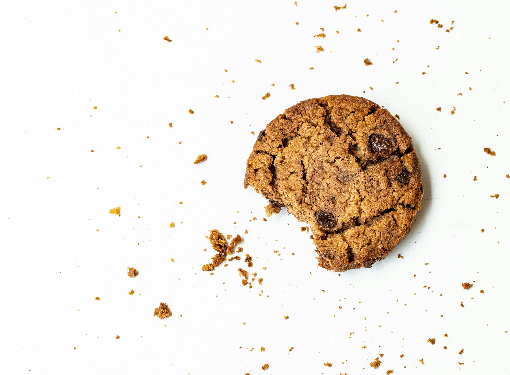 A chocolate chip cookie sitting on a worktop with a small bite out of it while there are crumbs surrounding the cookie itself