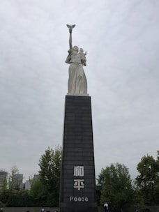 A tall statue stands atop a large pedestal, featuring a figure holding a dove aloft with one arm while supporting a child with the other arm. The pedestal displays the word 'Peace' and its translation in another language. Surrounding the monument are trees and some buildings are visible in the background under an overcast sky.